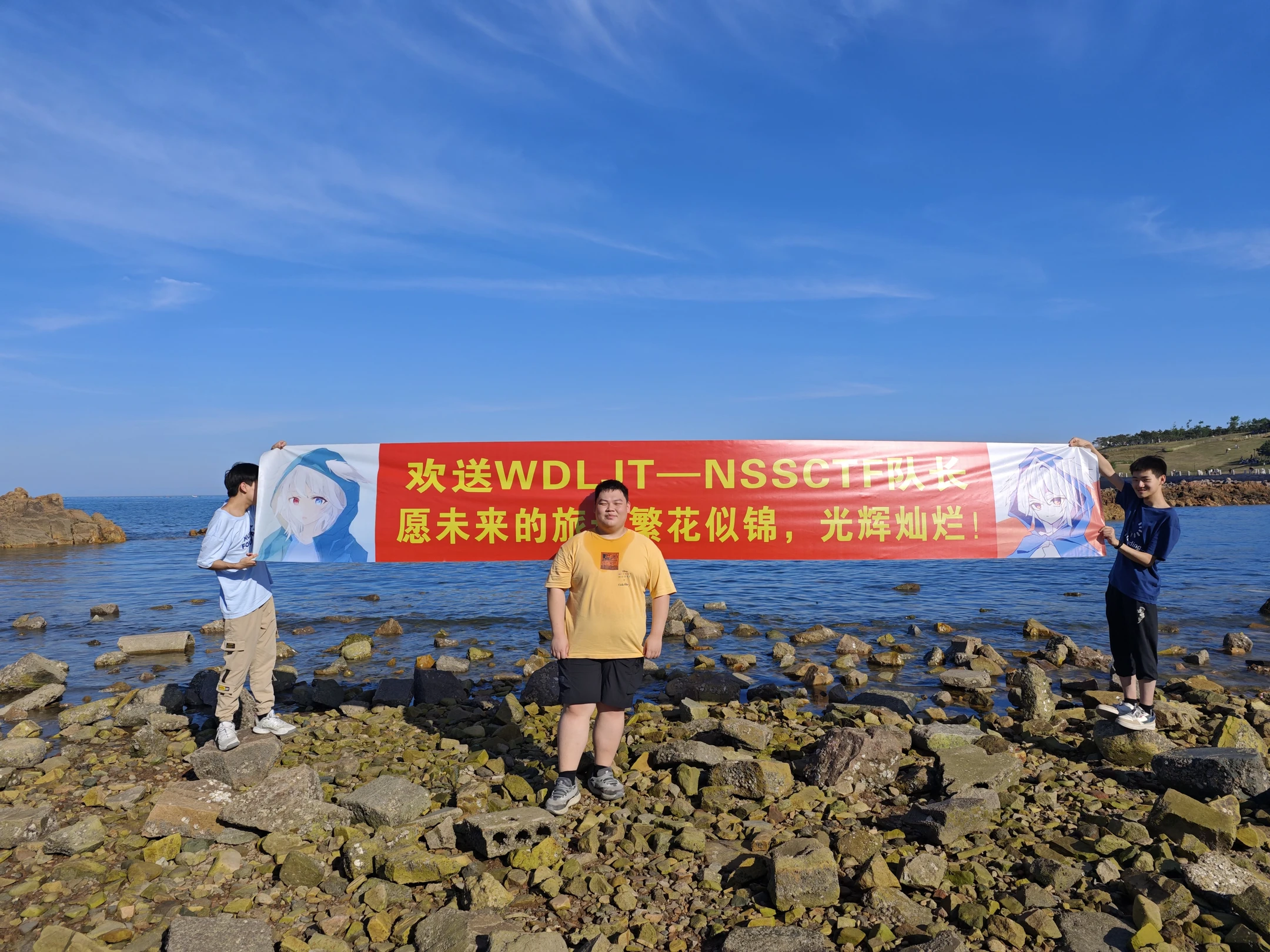 三位队员站在海边岩石上，举着写有欢送和祝福文字的横幅