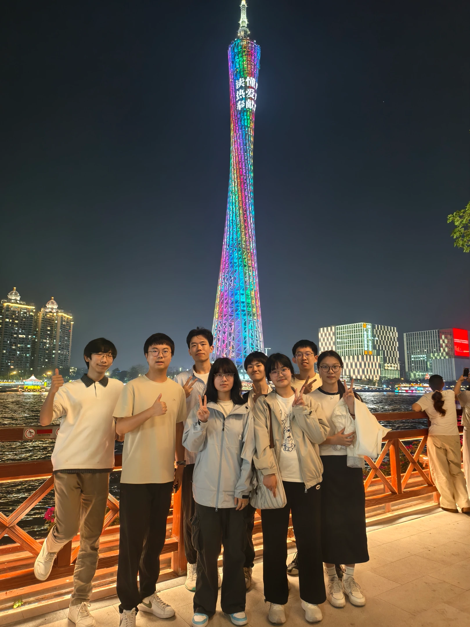 队员们站在江边栏杆前合影，身后是亮起彩色灯光的高塔